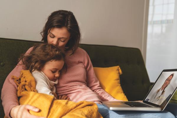 Mother comforting her young daughter, sitting on a couch with laptop showing a doctor on the screen.